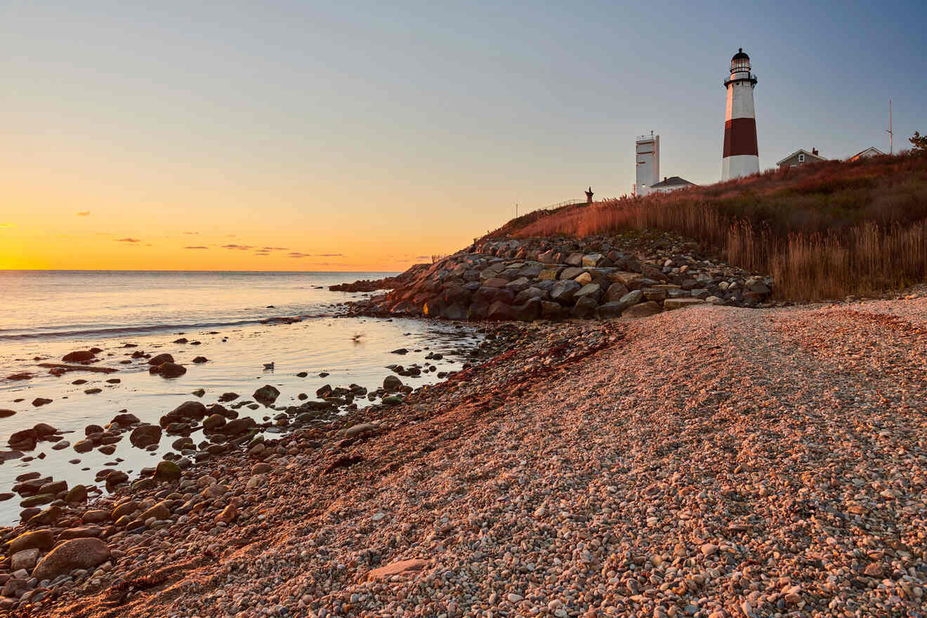 A historic lighthouse with red and white stripes stands on a grassy cliff, overlooking a rocky shore and calm sea under a blue sky with soft clouds