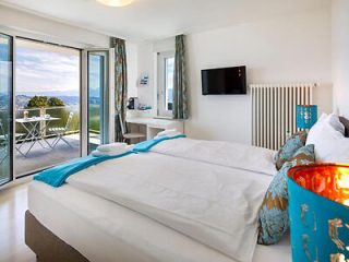Bright and airy hotel room with a double bed, crisp white linens, and turquoise accents, opening onto a balcony with a picturesque view.