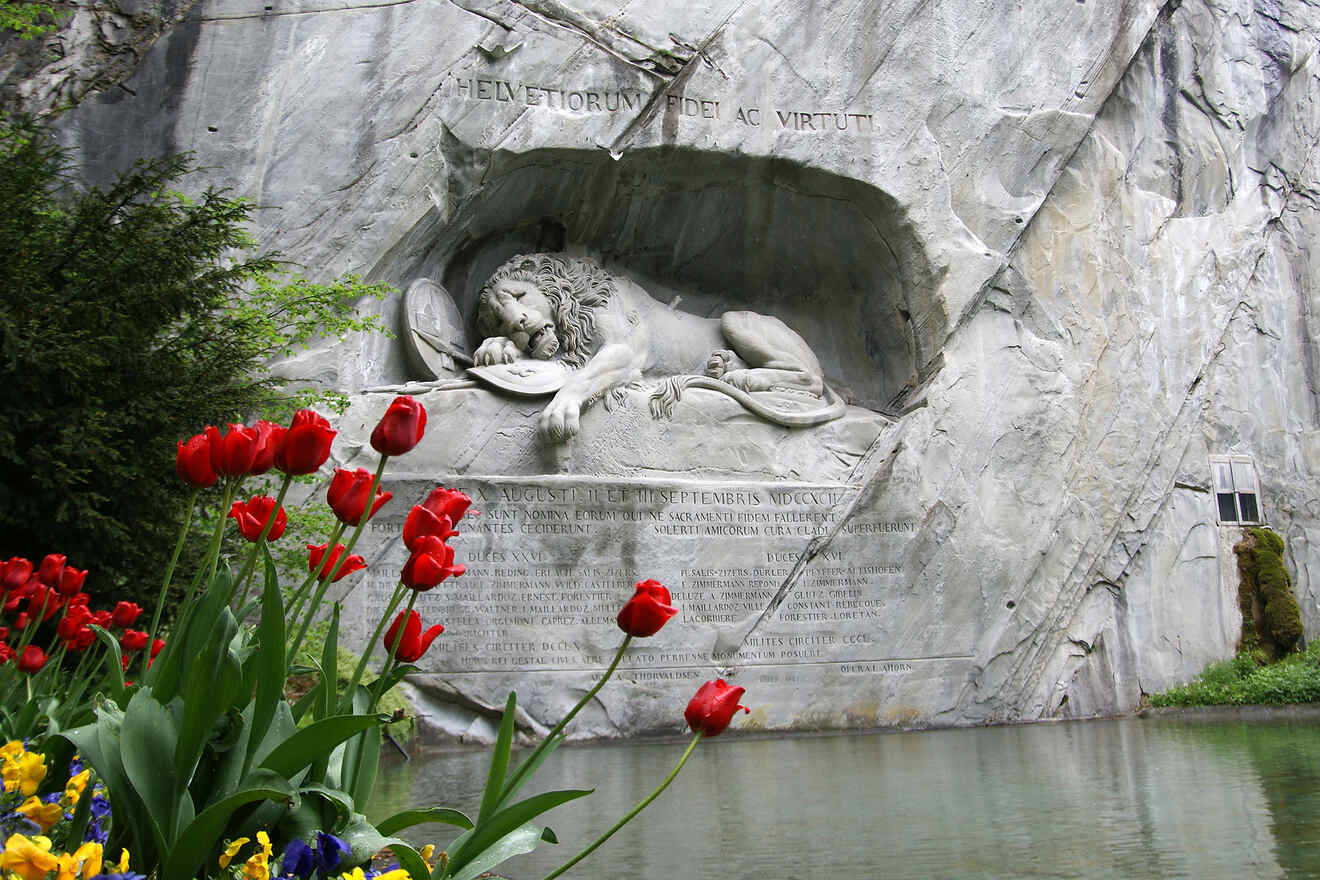 The Lion Monument in Lucerne, Switzerland, with a stone lion sculpture lying in a rock alcove above a pond, surrounded by blooming red tulips