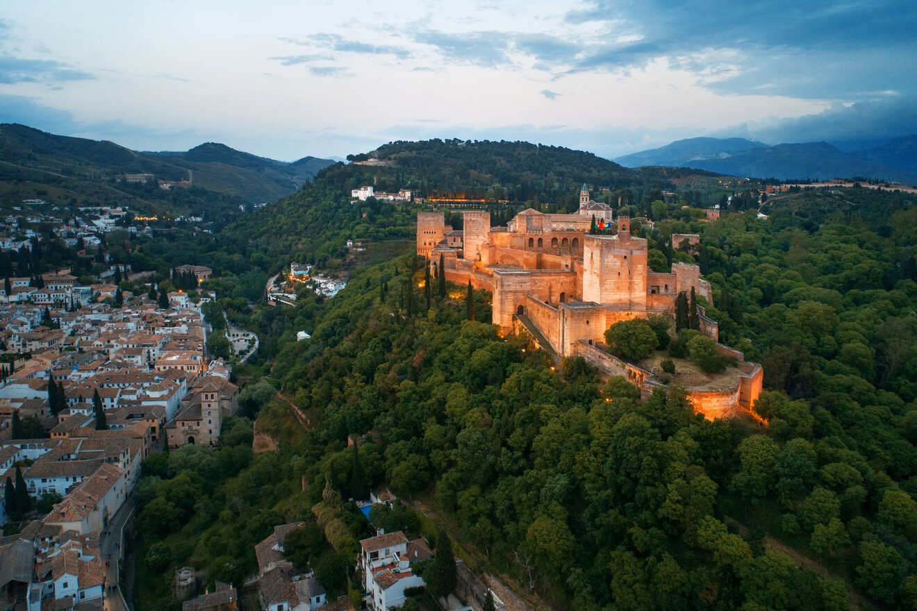 Aerial view of the Alhambra at twilight, its ancient walls lit against the lush hills of Granada and the fading light of the sky