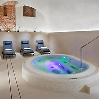 A serene indoor spa area with a circular hot tub surrounded by three reclined lounge chairs and lit by ambient lighting. The walls are white with exposed brick accents.
