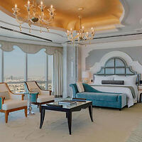 Luxurious hotel bedroom with a large bed, teal accents, and expansive windows offering a city view, under a glowing ornate ceiling.