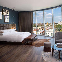Modern hotel room with panoramic windows overlooking a cityscape, featuring a minimalist design and neutral tones