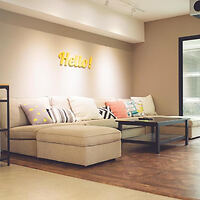 A beige sectional sofa with colorful pillows is placed against a wall with a "Hello!" sign. A black coffee table sits on a wooden floor. The room is modern with neutral tones and ample lighting.