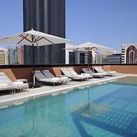 Rooftop hotel swimming pool lined with lounge chairs and umbrellas, overlooking a cityscape under a clear blue sky.