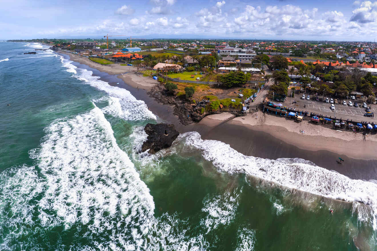 Aerial view of a bustling beachfront in Bali with waves crashing onto the shore, highlighting the contrast between the sandy beach and the lush greenery of the adjacent park and urban landscape