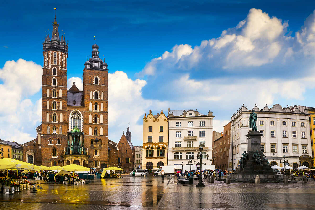 A historic square with a church featuring two towers, surrounding buildings with various architectural styles, yellow umbrellas from cafes, and a statue under a cloudy blue sky.