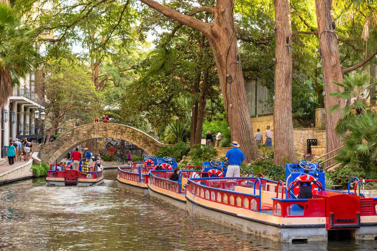 Tourists enjoying a guided boat tour on San Antonio Riverwalk, passing under a historic stone bridge surrounded by mature trees
