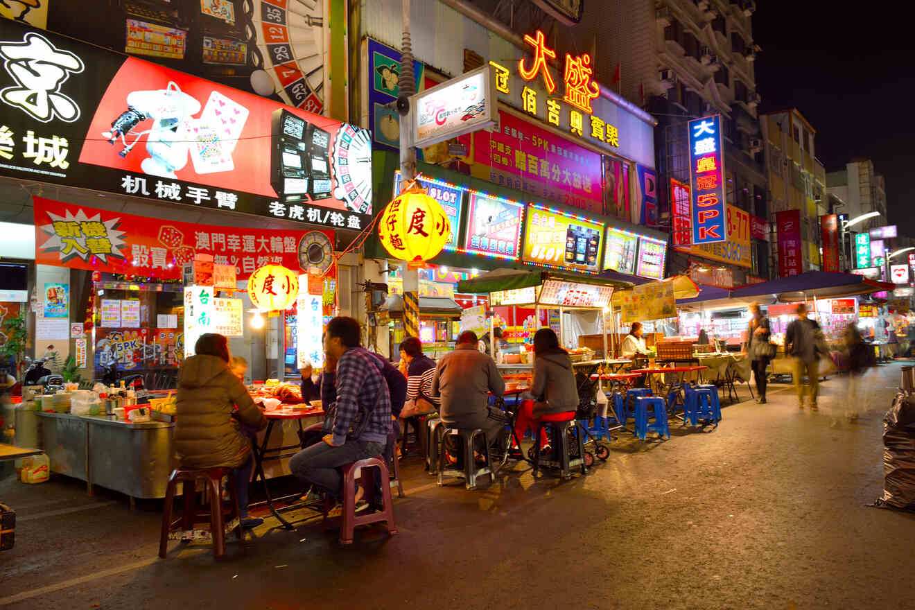 People sit on stools eating at food stalls amidst bright neon signs in a bustling night market.