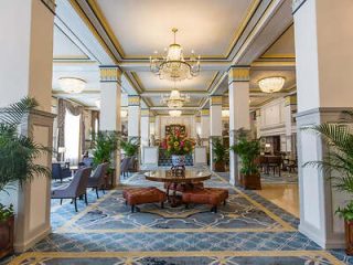 Opulent hotel lobby with classical architecture, featuring blue and white tiled flooring, a central floral arrangement, plush seating, and palatial columns.