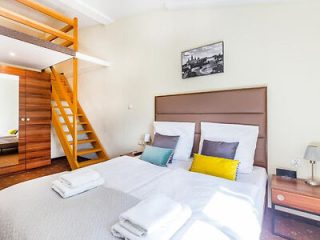 A well-lit bedroom with a double bed, two bedside tables, and a loft area accessible via wooden stairs. The bed has white linens and decorative pillows, with folded towels placed on top.