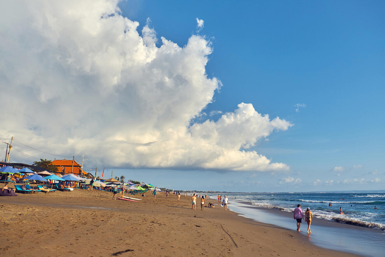 Beach scene with people strolling and sunbathing under a sky full of dramatic cumulus clouds, with traditional colorful umbrellas dotting the shoreline