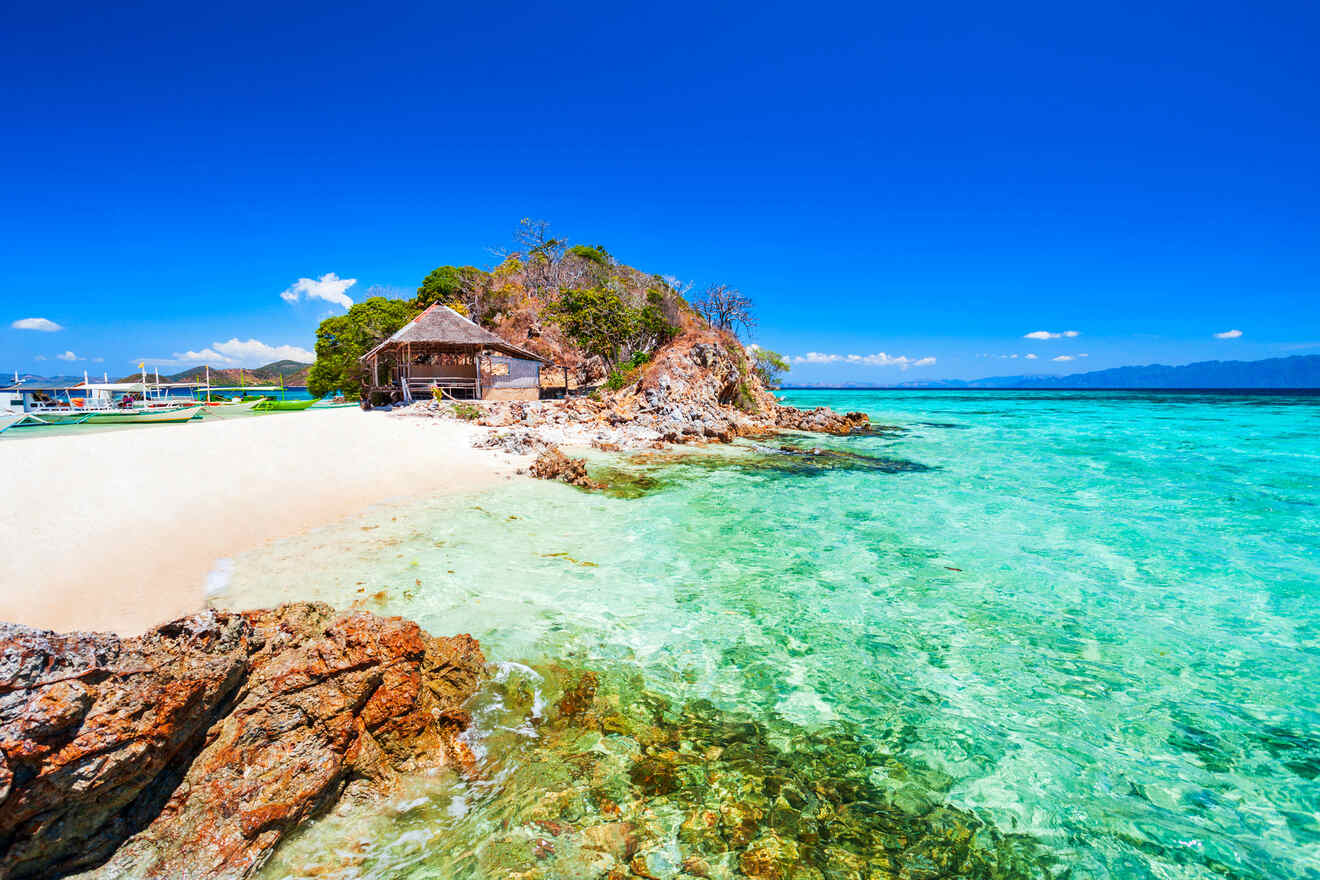 Clear turquoise water meets a white sandy beach next to a small hut on a rocky shore under a bright blue sky.