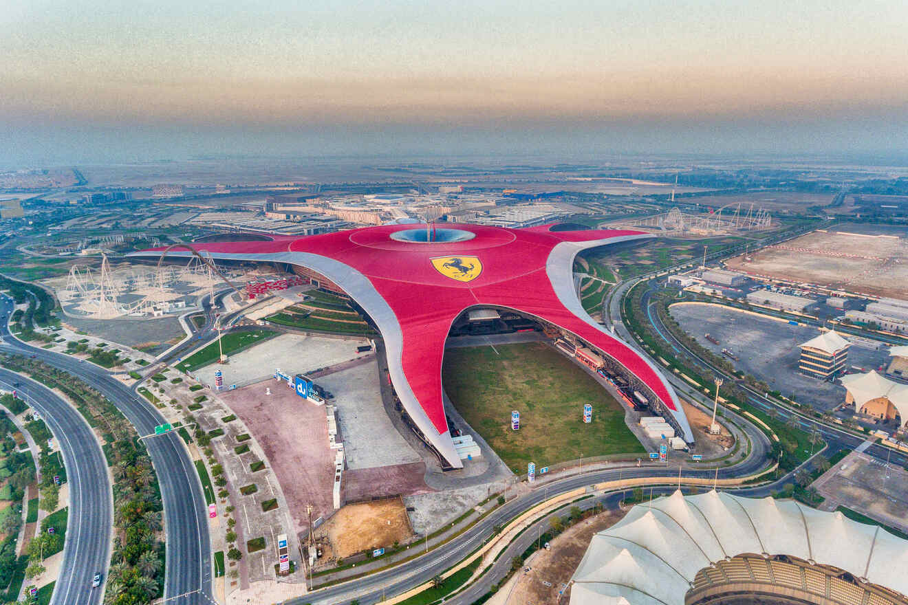 Aerial view of the iconic Ferrari World theme park on Yas Island, Abu Dhabi, with its distinctive red roof and Ferrari logo, surrounded by highways and the fading light of dusk.