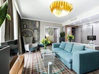 A modern living room features a blue sofa, glass coffee tables, a patterned wall, a large window with curtains, and a yellow chandelier overhead. The room is accented with plants and patterned rugs.