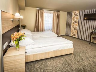 A hotel room with a double bed, soft lighting, a desk, a TV, and floral decorations. Beige and neutral tones dominate the room's decor, which includes a patterned wallpaper and curtains.