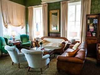 Traditional living room space with vintage charm, featuring a large white sofa, leather armchair, green upholstered chairs, and antique furniture against green patterned wallpaper.