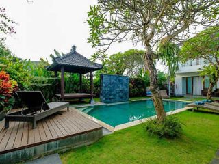 Private villa backyard with a wooden pergola next to a serene blue swimming pool, surrounded by tropical plants and a well-manicured lawn, suggesting a peaceful retreat