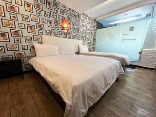 A bedroom features two double beds with white linens, a wood floor, and a wall decorated with numerous framed pictures. There is a glass wall partition leading to another room.