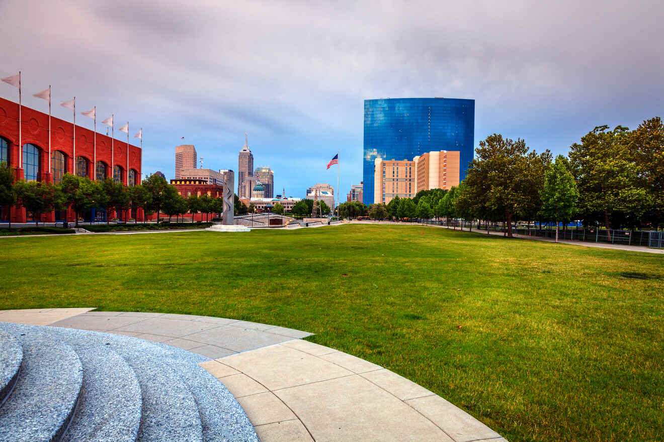 "Lush green park in Indianapolis with a modern sculpture in the foreground, the city’s skyline in the background, and a prominent blue glass building under a cloudy sky.