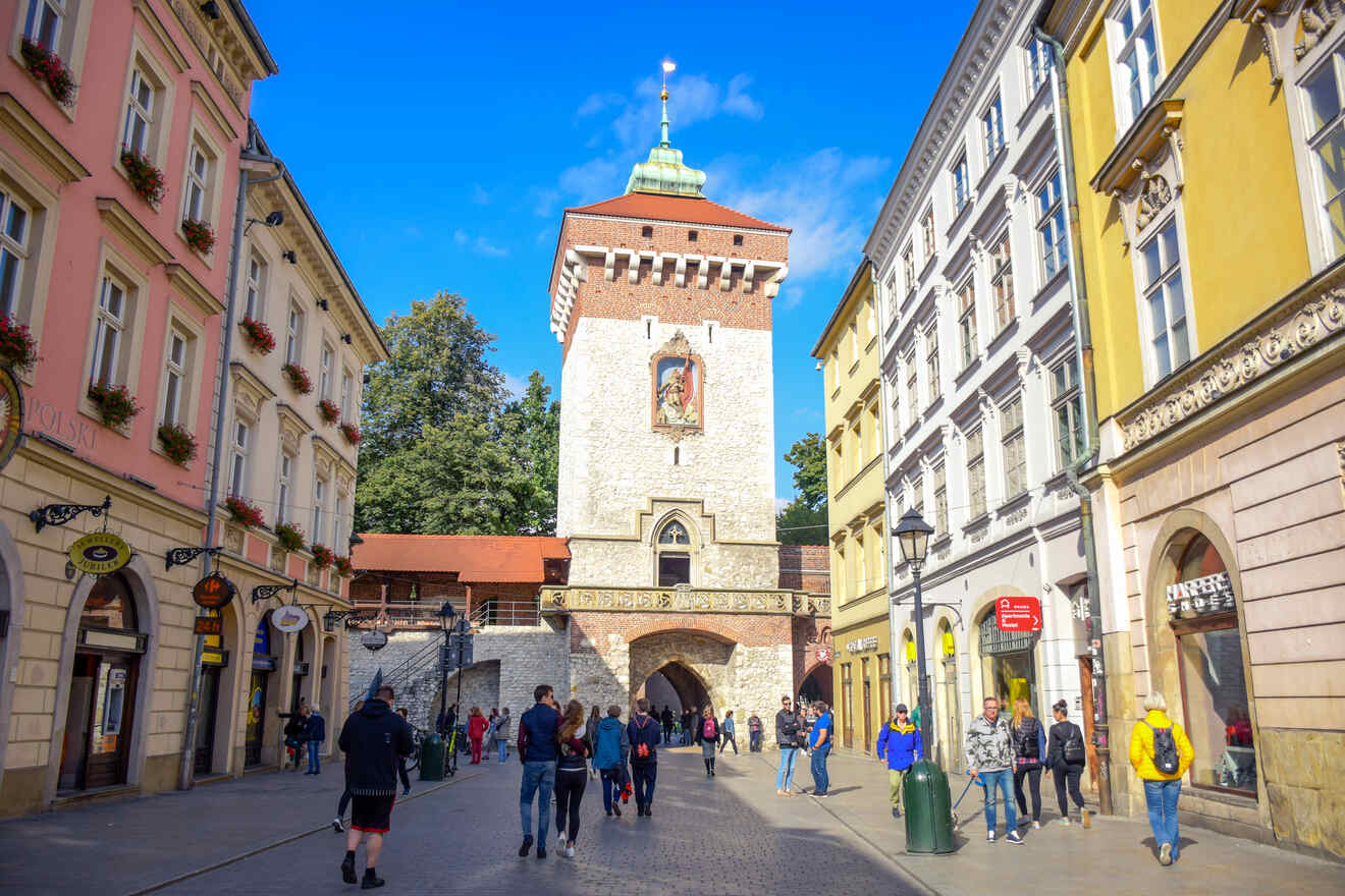 Street view of Saint Florian's Gate in Kraków, Poland, with people walking along a cobblestone road flanked by colorful historic buildings under a clear blue sky.