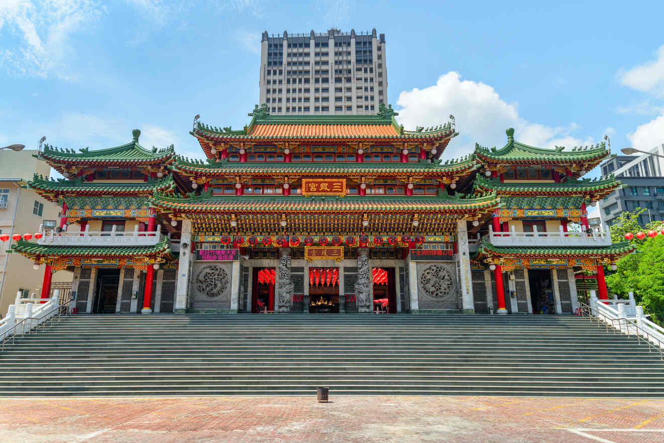 Front view of a traditional Chinese temple with ornate decorations, green-tiled roofs, and red lanterns. A modern high-rise building is visible in the background.