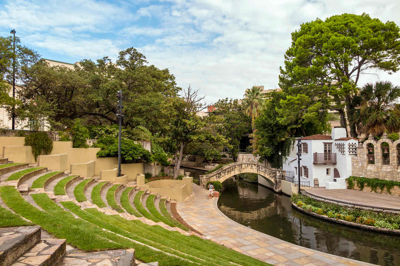 Amphitheater seating by the San Antonio Riverwalk with a serene waterway and a quaint stone bridge, surrounded by lush greenery and mature trees