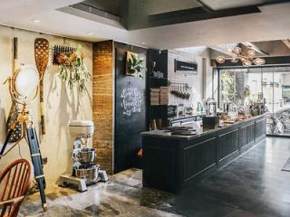 A cozy café interior featuring stylish decor with plants, hanging tools, and a chalkboard. The counter is equipped with coffee machines and accessories, while natural light filters through large windows.