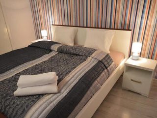 A neatly made bed with a patterned comforter and two pillows, flanked by two lit bedside lamps on white nightstands, against a striped wallpaper. Folded towels are placed on the bed.
