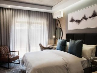 Elegant hotel room featuring a comfortable bed with dark headboard, blue accent pillows, a leather armchair, and minimalist artwork above the bed.