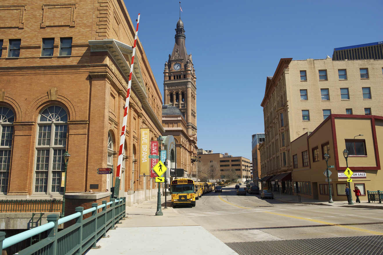 Street view of a historic clock tower rising above a city street lined with brick buildings and a 'Live Drama' theater sign in Milwaukee 