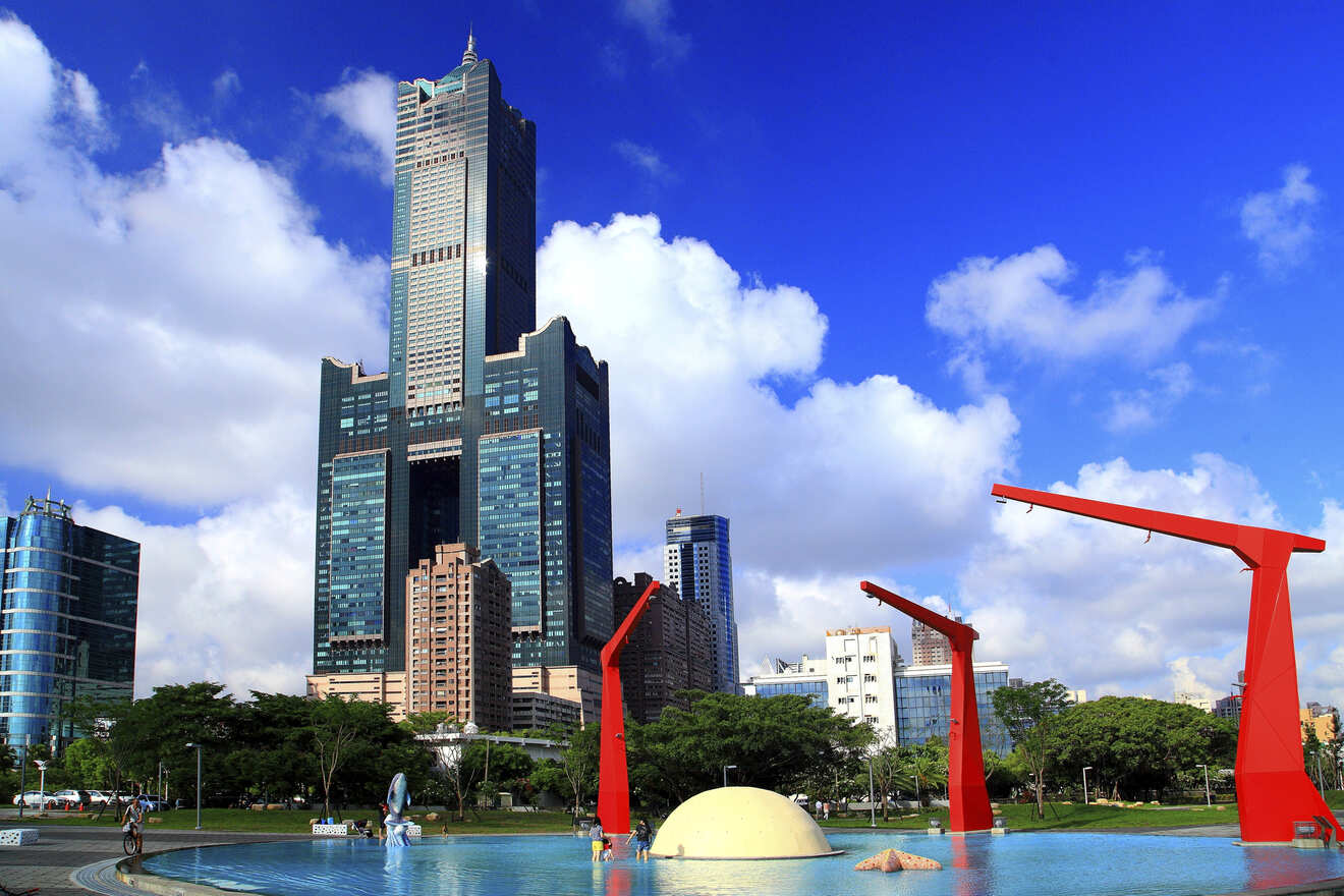 A tall skyscraper, the 85 Sky Tower, stands against a blue sky with scattered clouds, surrounded by smaller buildings and green trees. In the foreground, there is a water feature and red sculptures.