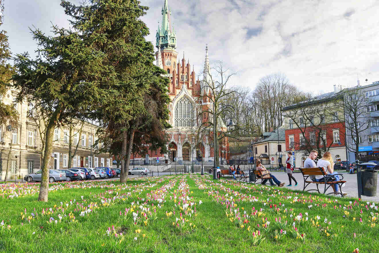 A Gothic-style church with a tall spire is situated behind a green lawn dotted with blooming flowers. People are sitting on benches along the path in front of the church. Buildings flank the church area.