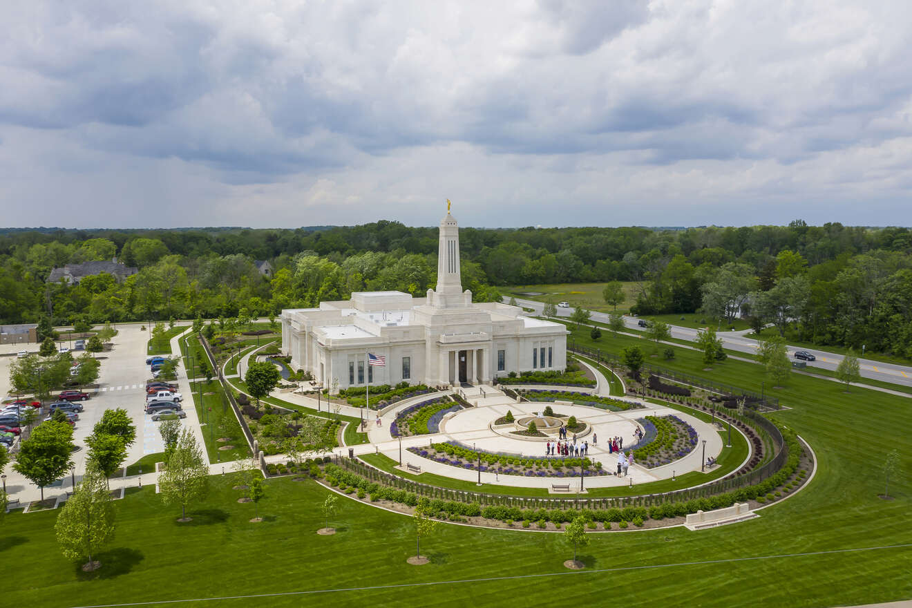 Aerial view of a white neoclassical temple-like building with a central spire, landscaped circular driveway, and green surroundings under a cloudy sky