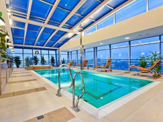 Indoor pool area with a glass ceiling and walls, several lounge chairs along the sides, and greenery accents. The pool has a stainless steel ladder and overlooks an exterior view through large windows.
