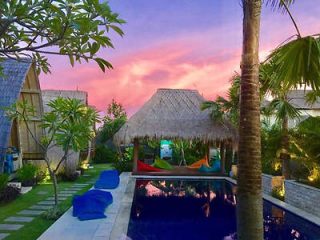 A tropical villa’s pool area at dusk, bathed in the pink hues of sunset, with a thatched-roof gazebo and lush greenery enhancing the peaceful retreat vibe.