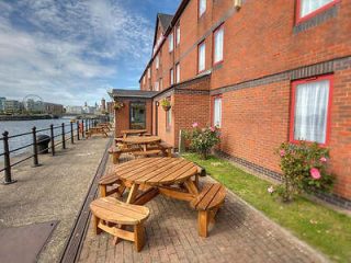 Waterfront outdoor seating area with wooden benches, overlooking a calm canal with red-brick buildings and a clear blue sky in the background