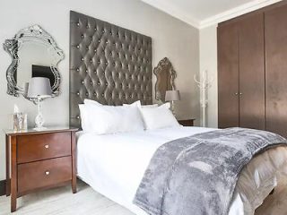 A luxurious hotel bedroom with a plush white bed, tufted gray headboard, mirrored furniture, and elegant decor.