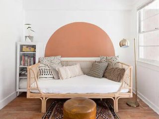 A chic nook featuring a rattan daybed with a half-moon painted wall overhead, eclectic cushions, a round leather pouf, and a floor lamp
