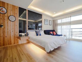 Modern bedroom with a double bed, wood-paneled accent wall, wall-mounted decor, a blue couch, large window with sheer drapes, and wooden flooring.