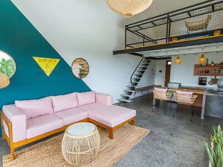 A chic, open-plan living room with a pink sofa and dining area, featuring a loft-style upper level and a vibrant blue accent wall with eclectic decor.
