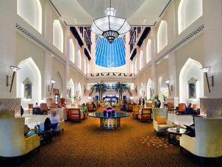 Opulent hotel lobby with high ceilings, ornate arches, and a large chandelier, filled with guests and seating areas.