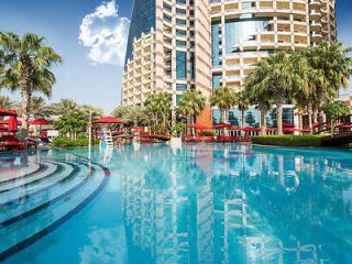 Resort-style pool with clear blue water, surrounded by palm trees and red poolside umbrellas against a backdrop of a high-rise building.