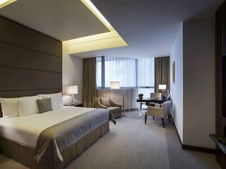 Contemporary hotel bedroom with a large bed, plush headboard, and a well-appointed work area, bathed in soft lighting.