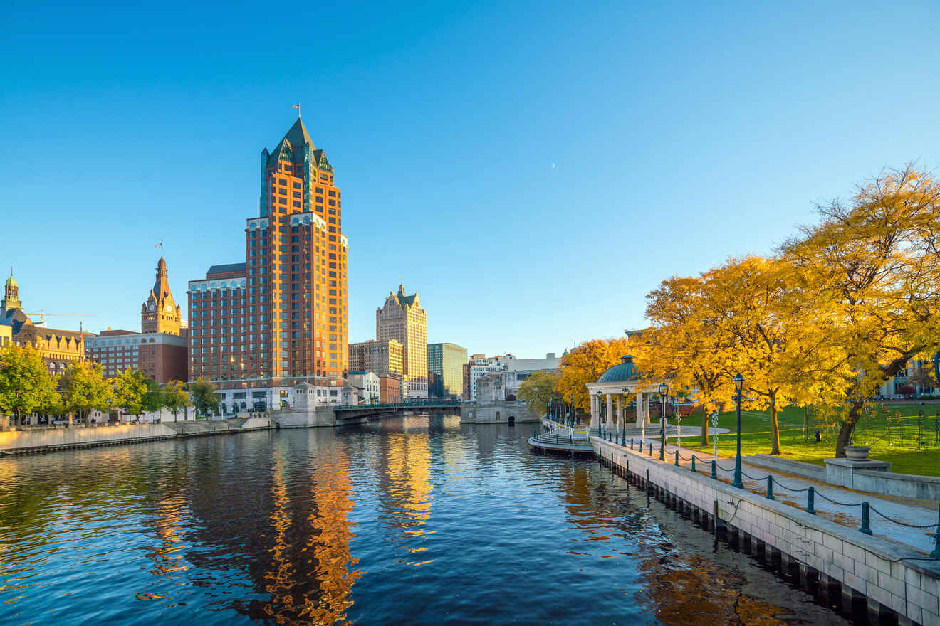Skyline of Milwaukee with autumnal trees, tall buildings reflected in the river, and a clear blue sky