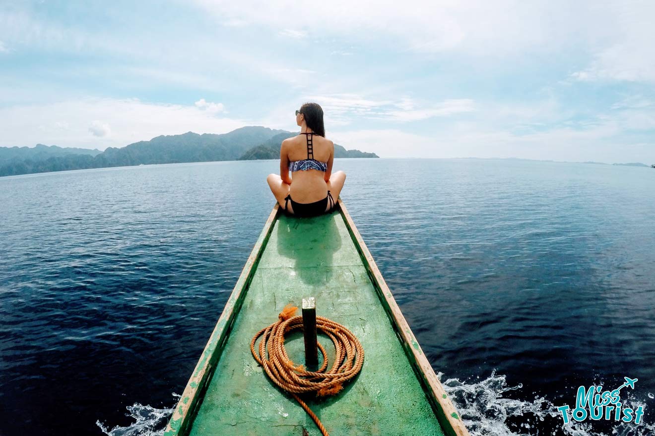 Back view of writer of the post sitting on the tip of a boat over calm water, facing distant mountains.