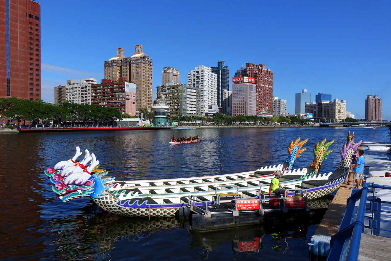 A decorative dragon boat is docked at a pier with a city skyline in the background on a sunny day. Another dragon boat is seen racing on the water.