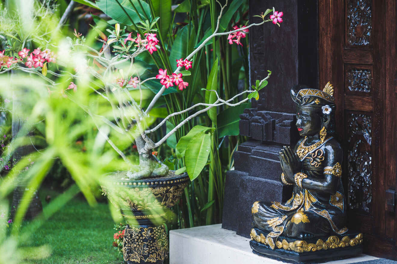 A traditional Balinese statue in prayer pose, adorned with intricate gold detail, set against a backdrop of tropical flora and architectural woodwork