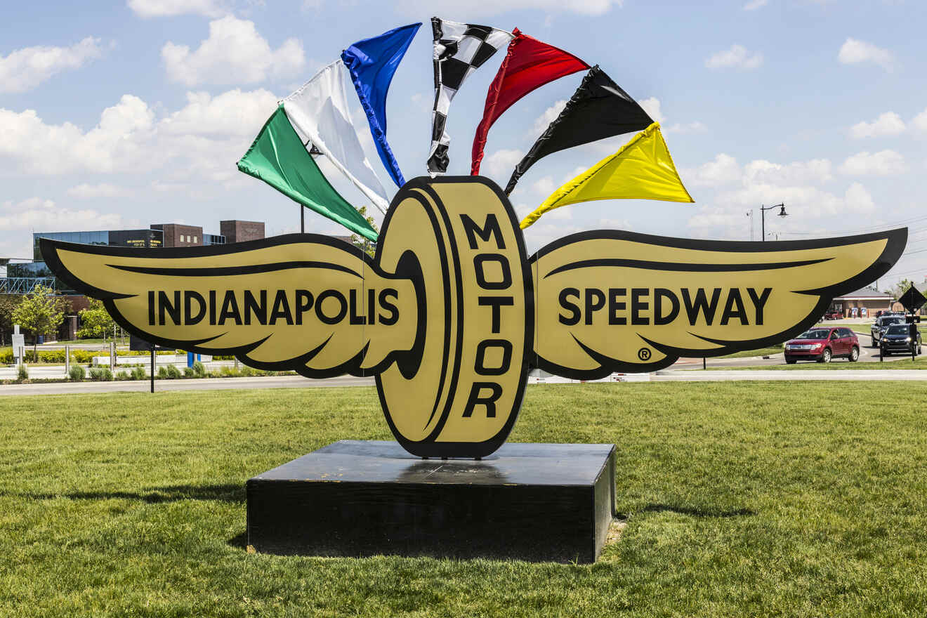 Iconic Indianapolis Motor Speedway logo with a wing and wheel, flanked by multicolored racing flags, set in a well-kept lawn area