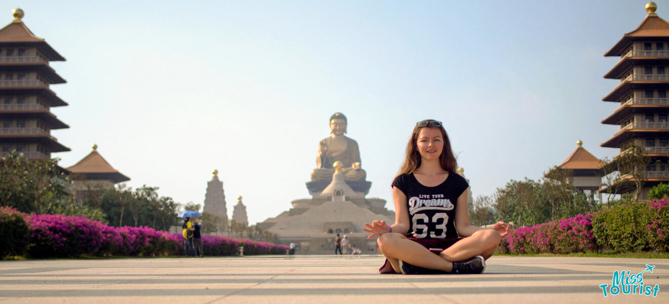 Author of the post sits cross-legged on a paved pathway with hands in a meditative pose, in front of a large statue of Buddha flanked by symmetrical temple structures.